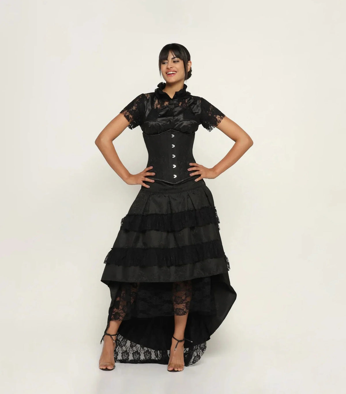 A woman modeling a black gothic-style dress with a lace corset bodice, short lace sleeves, a tiered ruffled skirt, and a high-low hemline, set against a plain white studio background.
