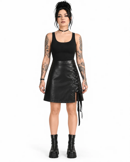 A model with tattoos stands against a white studio background wearing a black sleeveless top, a black faux-leather mini skirt with lace-up side detailing, and black platform ankle boots.