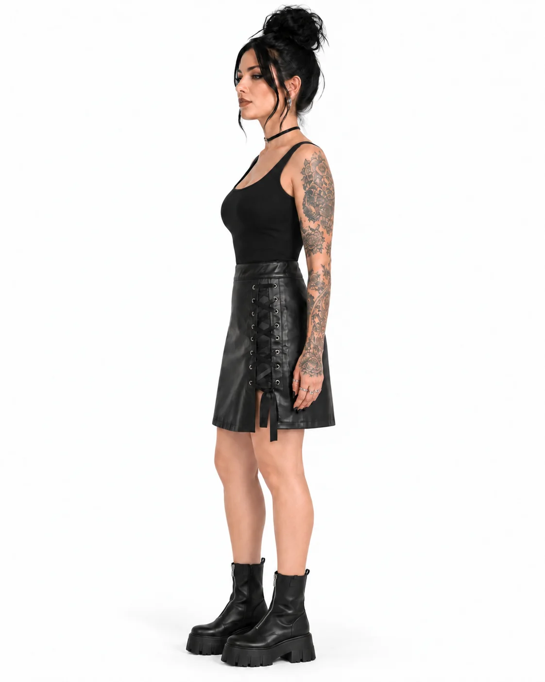 A woman with dark hair in a bun and visible arm tattoos stands in profile against a plain white studio background, wearing a black sleeveless top, a black faux-leather mini skirt with front lace-up detailing, and black chunky ankle boots.