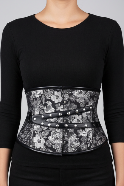 A black waist trainer corset belt with a black and white floral lace overlay and silver metal studs, worn by a model in a black long-sleeve top.