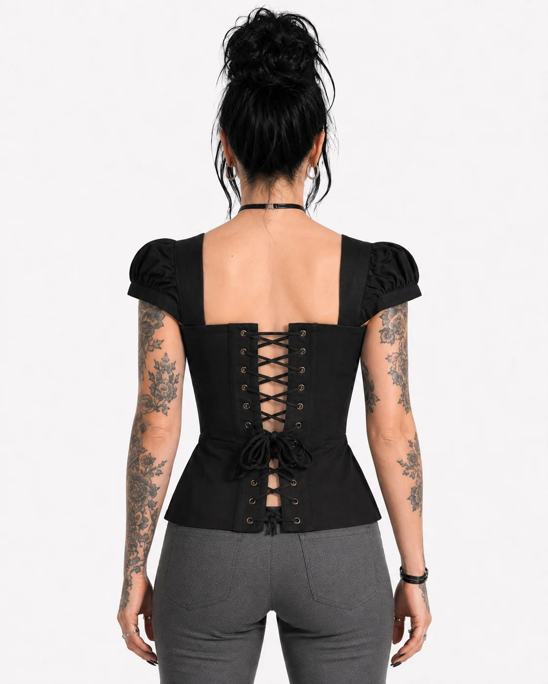 A woman viewed from the back wearing a black short-sleeve corset-style top with puff sleeves and a lace-up back, paired with gray pants, against a plain light background.