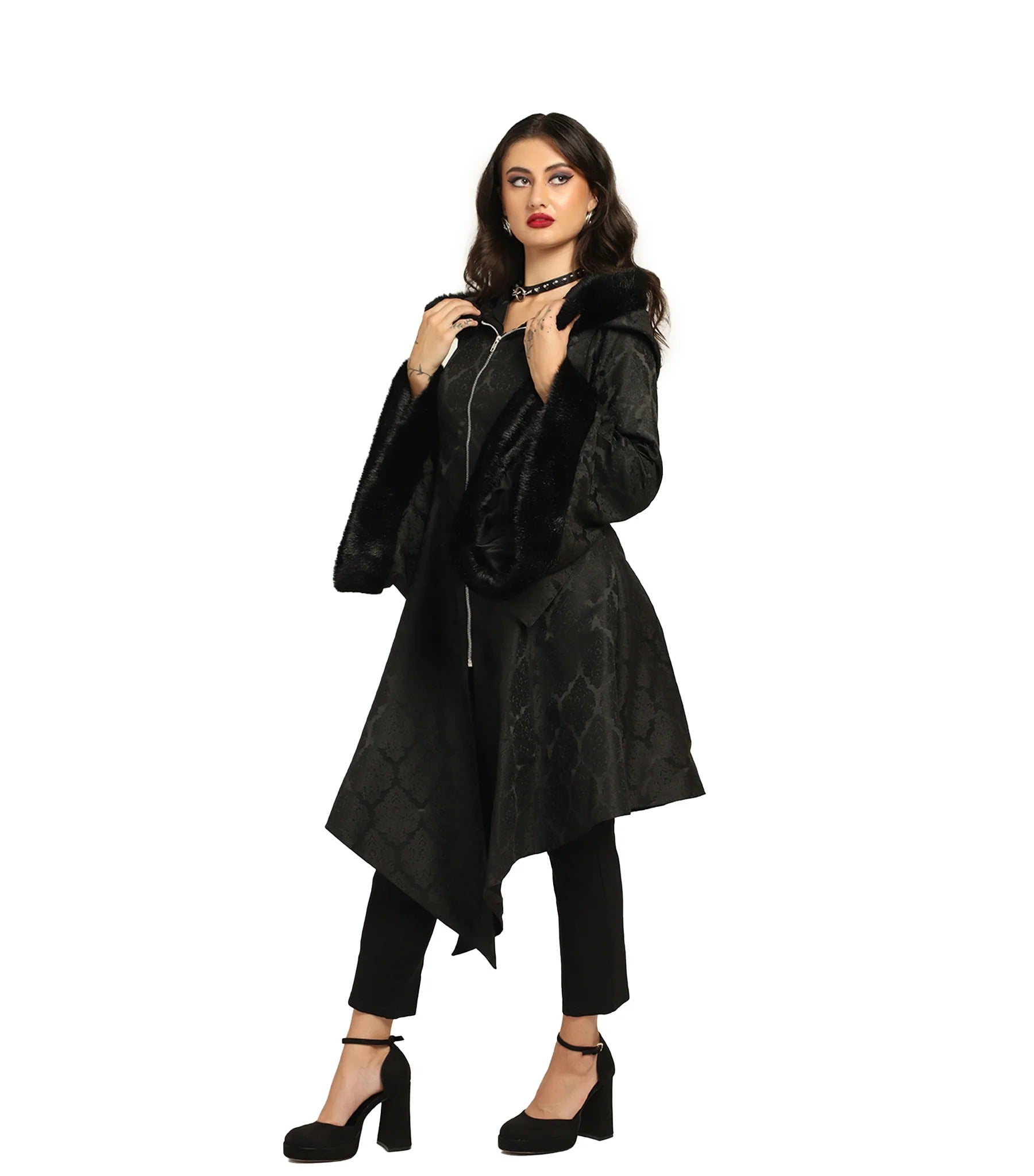 A woman modeling a black asymmetrical coat with faux fur trim and a snakeskin pattern, paired with black pants and heels, against a plain white background.